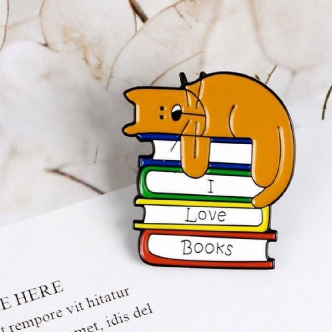 Orange cat resting on colourful books with “I Love Books” text enamel pin, approximately 1.1 × 0.9 inches.