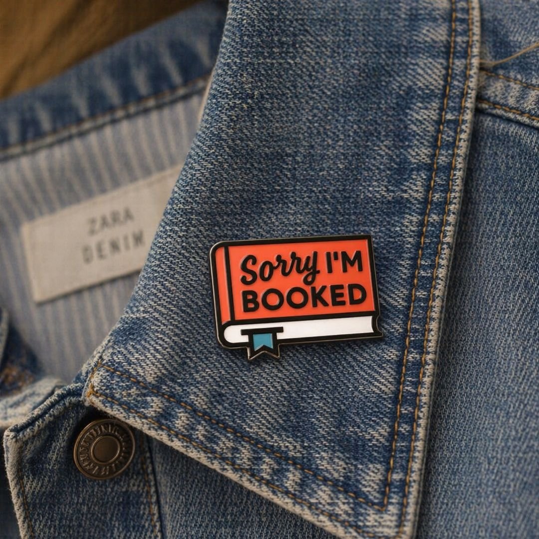 Sorry I'm Booked pin with orange lettering and black outline displayed on a blue denim jacket.