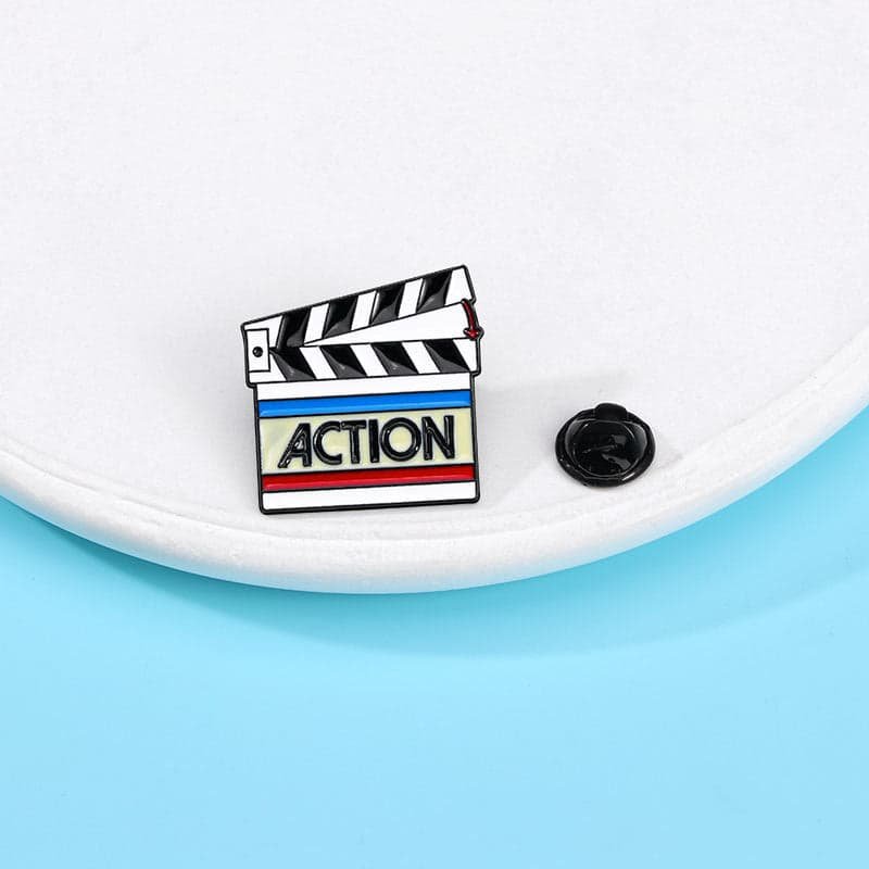 Action enamel pin featuring a black and white movie clapperboard with colourful stripes and ACTION lettering.