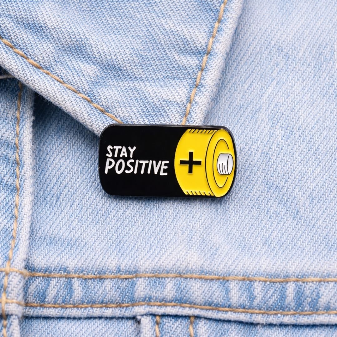 Stay Positive enamel pin in black and yellow battery design attached to a light blue denim jacket.