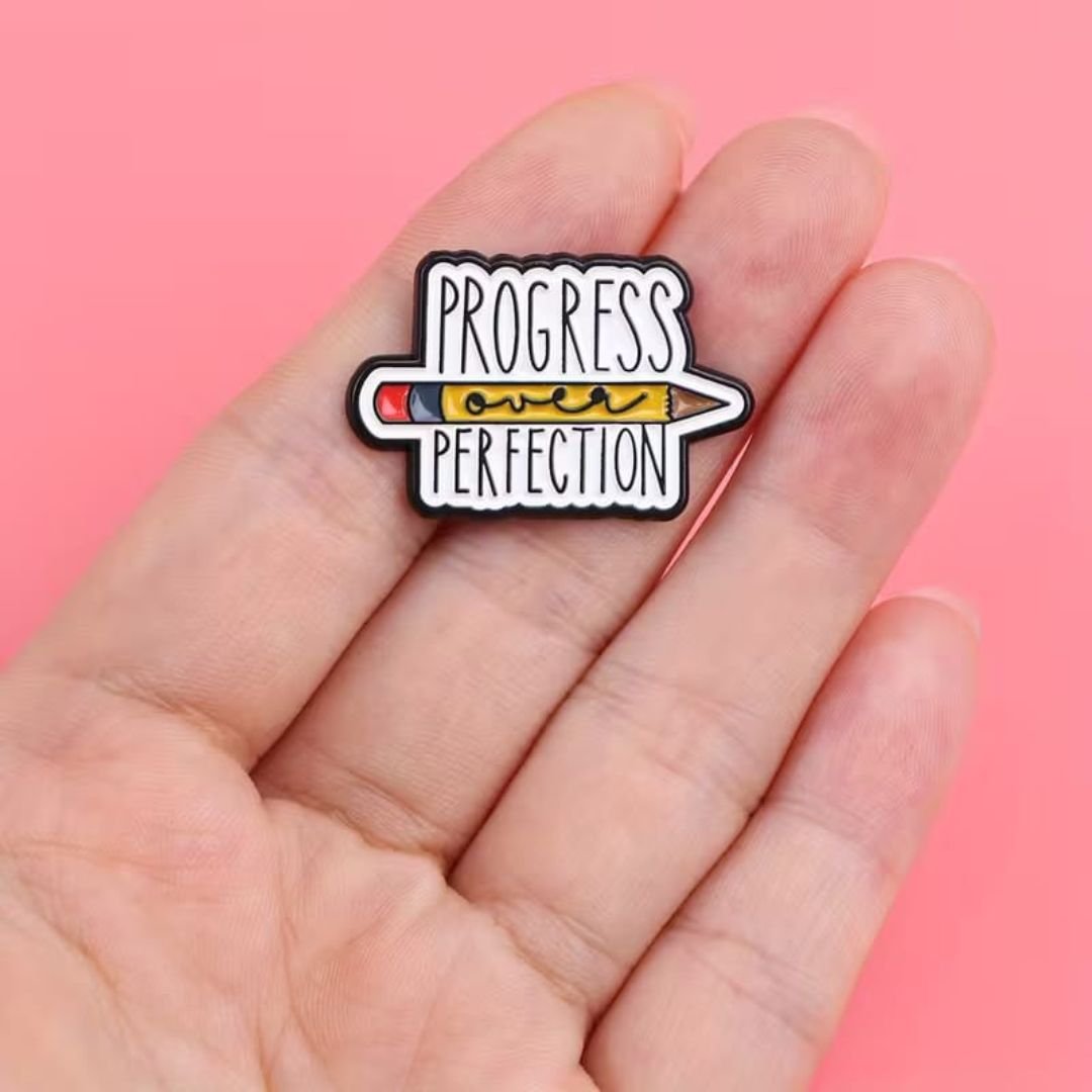 Progress Over Perfection enamel pin featuring a colourful horizontal pencil illustration.