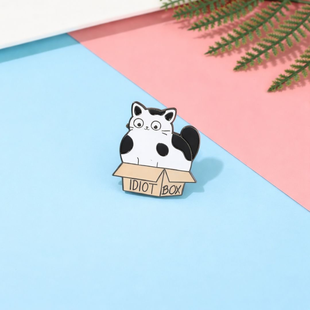 Idiot Box enamel pin featuring a black and white cat in a brown cardboard box.