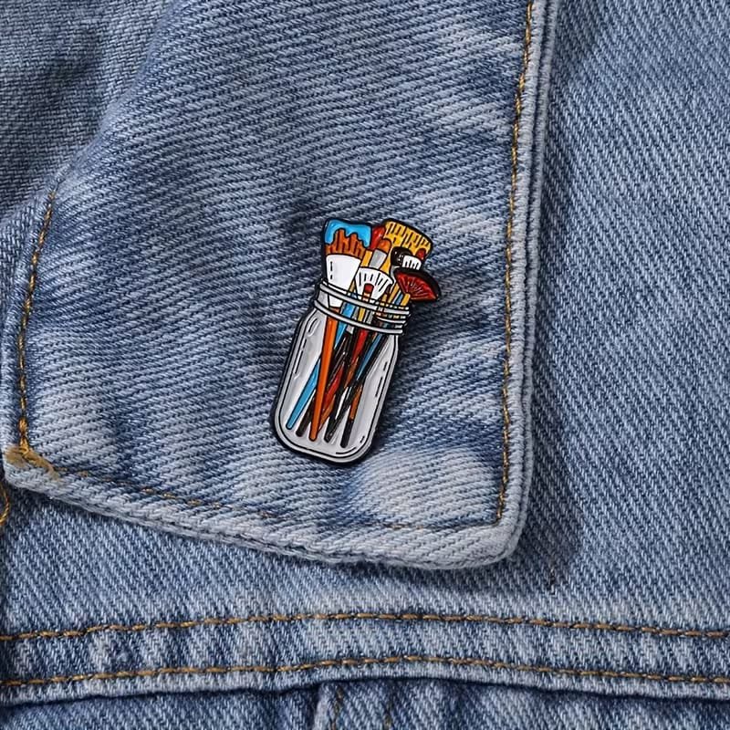 Paint brush jar enamel pin with colourful brushes, approximately 3.3 cm tall and 1.7 cm wide.
