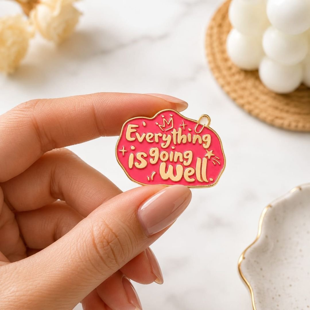 Pink and red Everything Is Going Well enamel pin with gold-tone outline held between fingers